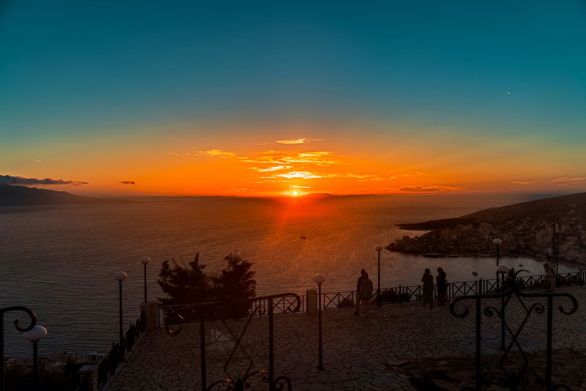 Sonnenuntergang über einer Bucht an der albanischen Küste, im Vordergrund eine Promenade mit schmiedeeisernem Geländer und Laternen